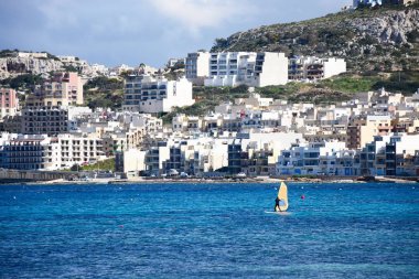 Windsurfer in the bay with town buildings to the rear, Mellieha, Malta. clipart