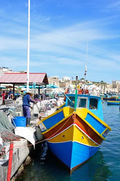 Tradiční maltský Dghajsa rybářské lodě v přístavu s domy na nábřeží na zadní a místní muž třídění své rybářské sítě na nábřeží, Marsaxlokk, Malta. — Stock fotografie