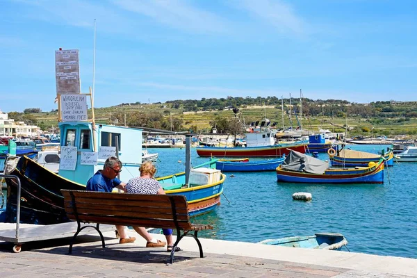 Tradiční maltský Dghajsa rybářské lodi v přístavu s pár sedí na lavičce v popředí, Marsaxlokk, Malta. — Stock fotografie