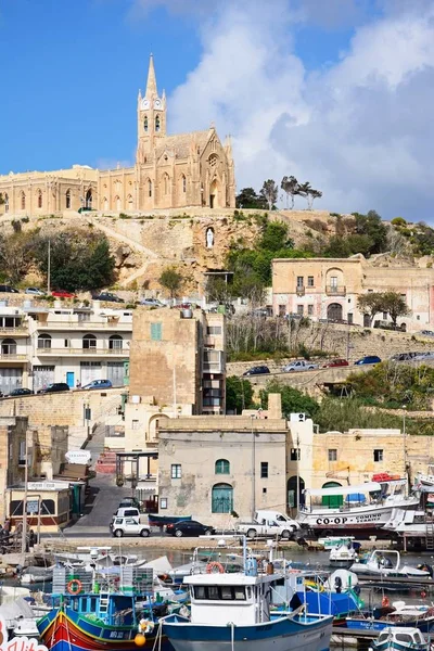 Traditionella fiskebåtarna i hamnen med vår fru av Lourdes kyrkan på bergssluttningen till baksidan, Mgarr, Gozo, Malta. — Stockfoto