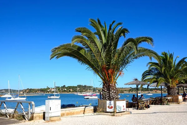 Turisté relaxační v kavárně chodníku podél promenády s výhledem na jachty kotvící v ústí, Alvor, Algarve, Portugalsko. — Stock fotografie