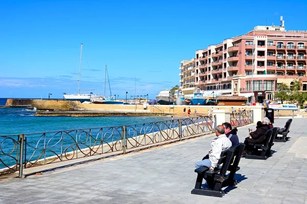 Tourists sitting on benches along the promenade with views towards beach and harbour, Marsalforn, Gozo, Malta. — Stock Photo, Image