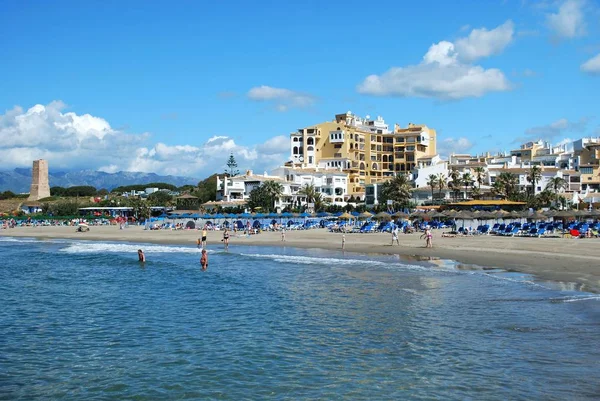 Turistas relajándose en la playa con una antigua torre de vigilancia y apartamentos en la parte trasera, Cabopino, Marbella, España . — Foto de Stock