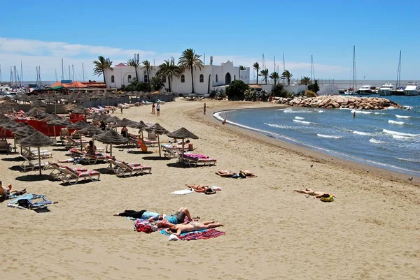 Turyści relaksujący się na plaży Daitona z przystanią na tyłach, Marbella, Hiszpania. — Zdjęcie stockowe