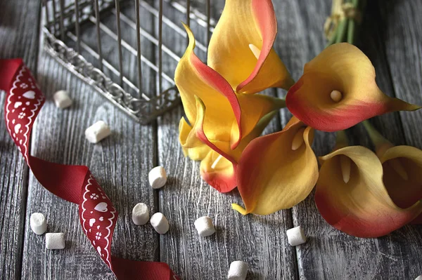 Orange gelbe Calla-Lilien mit roter Schleife und weißen Marshmallows auf grauem Holzgrund. — Stockfoto