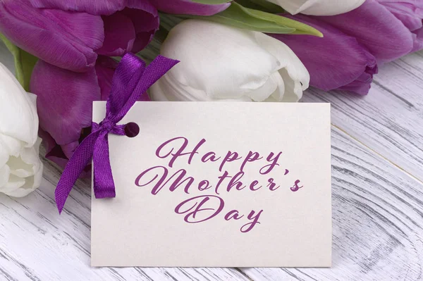 Tulipas roxas e brancas com papel branco em um fundo de madeira branco e lettering cartão feliz Dia das Mães . — Fotografia de Stock
