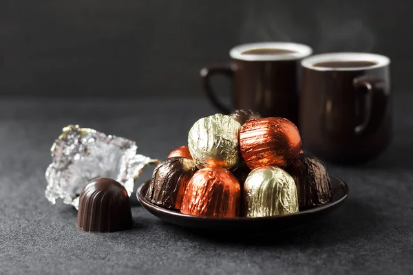 Шоколадні цукерки, загорнуті в різнокольорову фольгу і дві чашки кави на чорному фоні — стокове фото