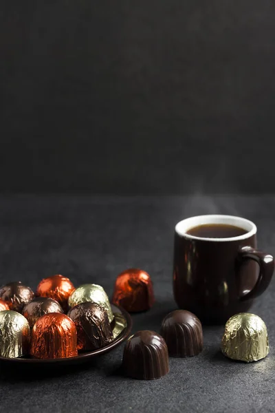 Шоколадні цукерки, загорнуті в різнокольорову фольгу і чашку кави на чорному тлі — стокове фото