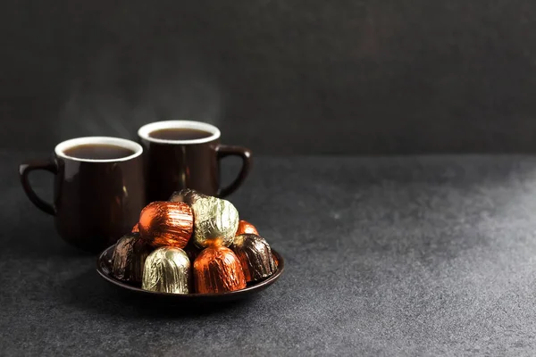 Permen coklat dibungkus dengan foil berwarna-warni dan dua cangkir kopi dengan latar belakang hitam — Stok Foto