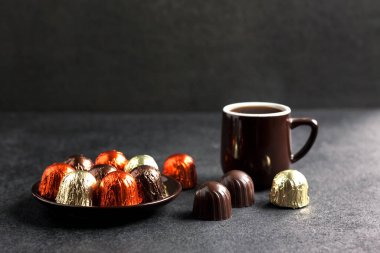 Bir tabakta çok renkli folyoya sarılı çikolatalar ve siyah arkaplanda fotokopi alanı olan bir fincan sıcak kahve