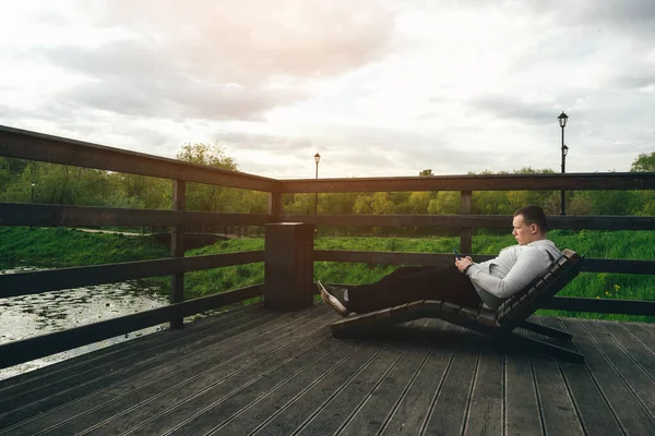 the guy sits on a bench bench in the park, and looks at the phone. enjoys nature. a tourist. sitting near the lake