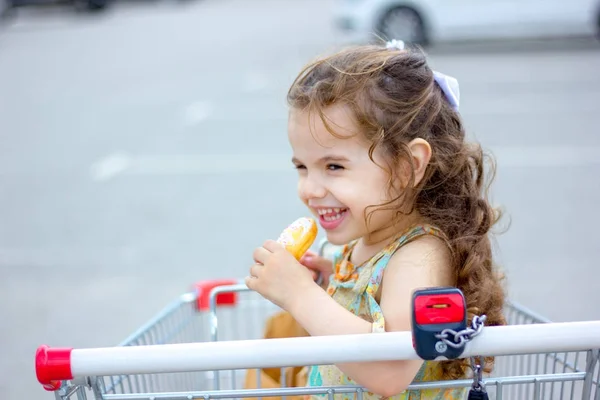 Niña sonriendo mientras come donut en el carrito de la compra en el estacionamiento del centro comercial. — Foto de Stock