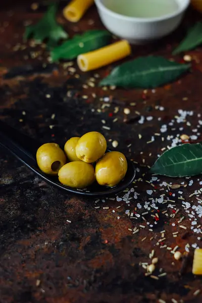 Olivy v černé lžíci obklopené dalšími ingrediencemi — Stock fotografie