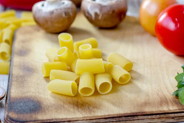Сухая паста ригатони на деревянной доске в окружении овощей. — стоковое фото