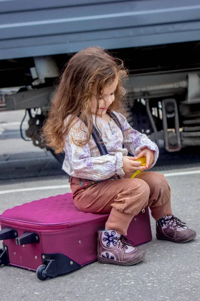 Niña sentada en la maleta en la estación de tren con el tren en el fondo . — Foto de Stock