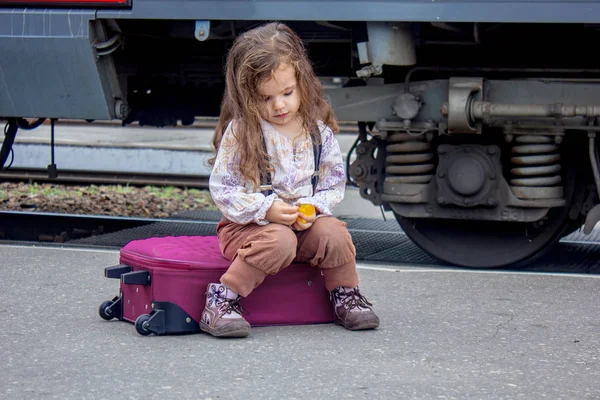 Niña sentada en la maleta en la estación de tren con el tren en el fondo . — Foto de Stock