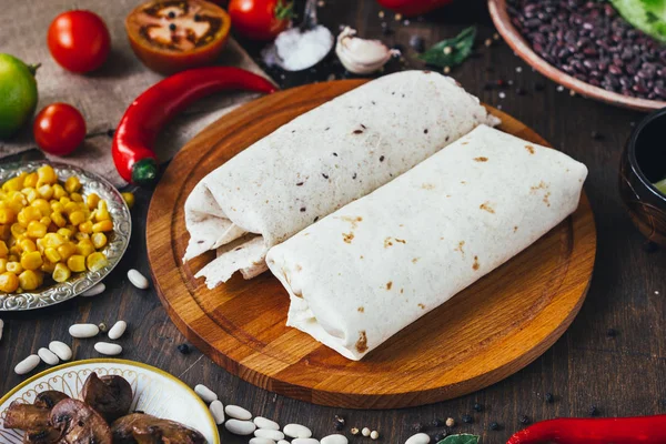 Vegetariánské burrito na dřevěné desce nad černým stolem obklopeno ingrediencemi. — Stock fotografie