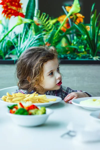 Niña linda sentada en un restaurante y comiendo papas fritas. — Foto de Stock