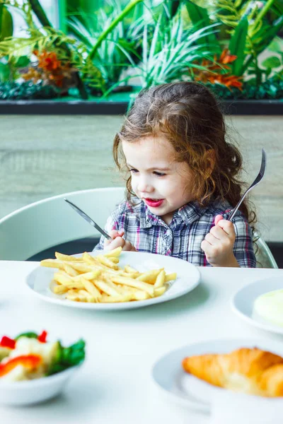 Niña almorzando en el restaurante con el cuchillo de mesa y tenedor en las manos. — Foto de Stock