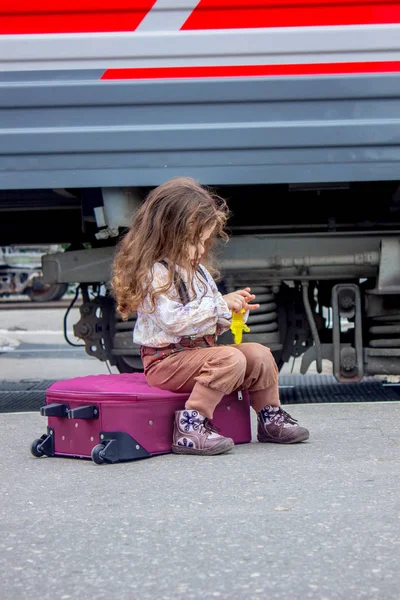 Niña sentada en la maleta en la estación de tren con el tren en el fondo . — Foto de Stock