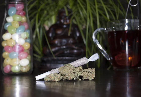 Pěkné closeup válcované marihuany plevel kloubu a pupeny, na dřevěné pozadí, soška Buddhy, barevné jellybeans a šálek čaje rooibos — Stock fotografie