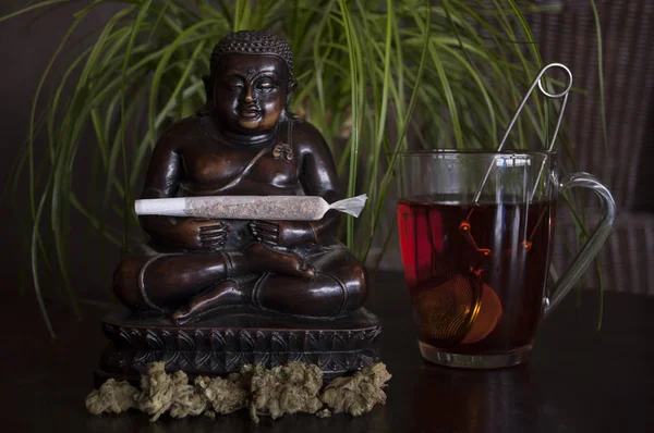 Joli gros plan de joint de marijuana roulée et bourgeons avec statuette de Bouddha et tasse de thé rooibos sur sol en bois — Photo