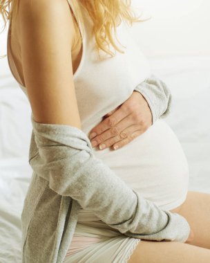 Beyaz tişörtlü sarışın hamile bir kadın yatakta oturuyor. Yüzü görünmüyor..