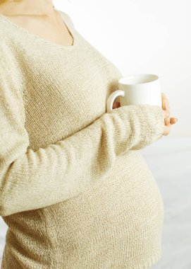 Süveter giymiş hamile bir kadın beyaz bir bardak tutuyor. Yüzü yok