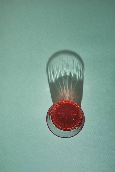 A glass of red juice casts a long shadow on a light green background.