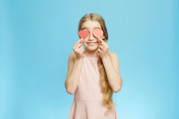 Isolierte kleine Mädchen bedeckt ihre Augen mit Papierherzen auf blauem Hintergrund. Einzelnes Halblanges Porträt. — Stockfoto
