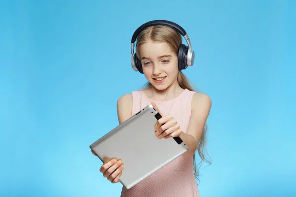 Güzel, küçük, neşeli kız tablette oyun oynuyor. Kız kulaklık takıyor. Mavi arkaplanda bir portre.