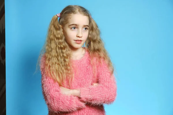 Ein kleines Mädchen im rosafarbenen Pullover steht vor blauem Hintergrund. Mädchen mit langen blonden Haaren — Stockfoto