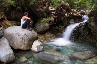 Latince bir orta yaş adam rahat bir pozisyonda akan bir su düşüş yanında bir kayaya oturuyor. Aslanlar doruklarına aslanlar Bay, Kuzey Vancouver, Bc, Kanada üst için yürüyüşe geçen resim.