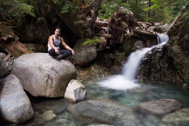 Latince bir orta yaş adam rahat bir pozisyonda akan bir su düşüş yanında bir kayaya oturuyor. Aslanlar doruklarına aslanlar Bay, Kuzey Vancouver, Bc, Kanada üst için yürüyüşe geçen resim.