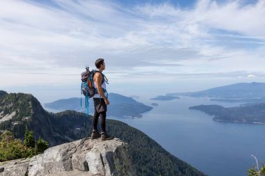 Latin American Male Hiker is standing on top of the rocky peak overlooking the beautiful landscape. Picture taken on the way up to The Lions Mountain, North of Vancouver, British Columbia, Canada. clipart