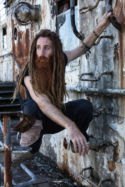 A rugged man with long beard and dreadlocks standing on an abandoned boat with rusty metal walls.