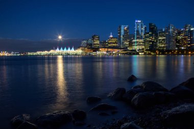 Beautiful view of Downtown Vancouver Skyline during the night with the full moon rising. Taken in Stanley Park around Coal Harbour, British Columbia, Canada. clipart