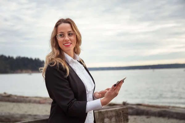 Young European Business Woman well dressed is holding her cell phone and smiling on a sandy beach. Picture taken West Vancouver, British Columbia, Canada.