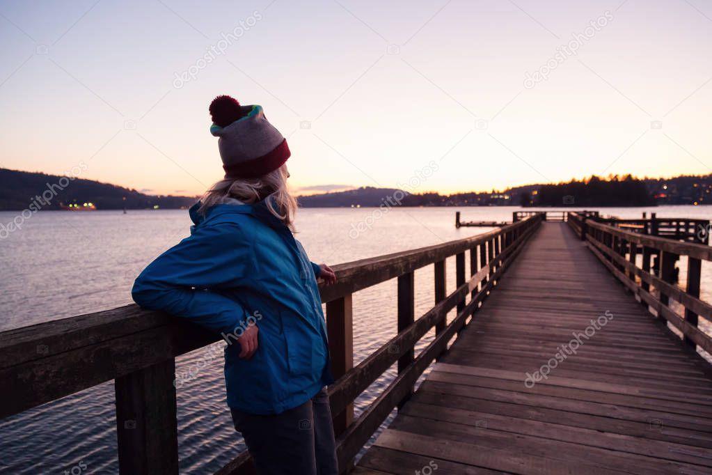 Young woman standing at a wooden quay in a park watching the beautiful scenery during a vibrant sunset.  Taken in Belcarra, Vancouver, British Columbia, Canada.