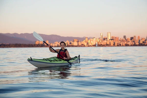 Beautiful woman sea kayaking in the ocean during a colorful and vibrant sunset with the city skyline in the backgroun. Taken in Jericho, Vancouver, British Columbia, Canada.