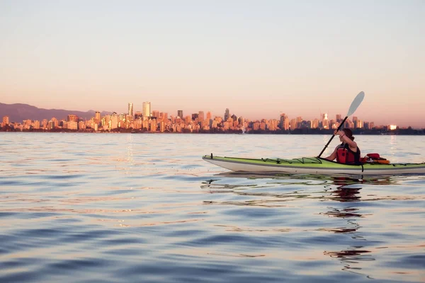 Beautiful woman sea kayaking in the ocean during a colorful and vibrant sunset with the city skyline in the backgroun. Taken in Jericho, Vancouver, British Columbia, Canada.