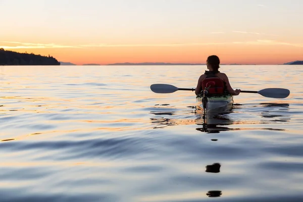 Woman on a sea kayak is paddling in the ocean during a colorful and vibrant sunset. Taken in Jericho, Vancouver, British Columbia, Canada.