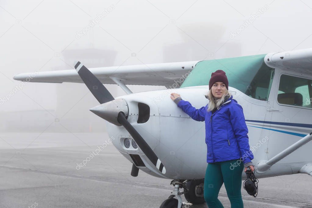 Young Caucasian Female Student Pilot is standing in front of a Single Engine Airplane at the Airport. Taken during a foggy winter morning in Pitt Meadows, Vancouver, BC, Canada.
