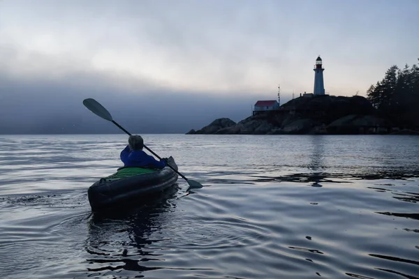 Adventurous woman is sea kayaking near a lighthouse during a vibrant and foggy winter sunset. Taken in Horseshoe Bay, West Vancouver, BC, Canada. Concept: adventure, holiday, vacation, explore