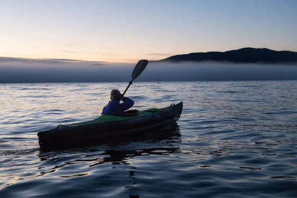 Adventurous woman is sea kayaking on an inflatable kayak during a vibrant winter sunset. Taken in Horseshoe Bay, West Vancouver, BC, Canada. Concept: adventure, holiday, vacation, explore