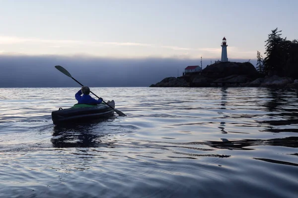 Adventurous woman is sea kayaking near a lighthouse during a vibrant and foggy winter sunset. Taken in Horseshoe Bay, West Vancouver, BC, Canada. Concept: adventure, holiday, vacation, explore