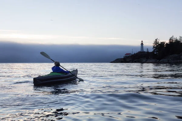 Adventurous woman is sea kayaking near a lighthouse during a vibrant and foggy winter sunset. Taken in Horseshoe Bay, West Vancouver, BC, Canada. Concept: adventure, holiday, vacation, explore