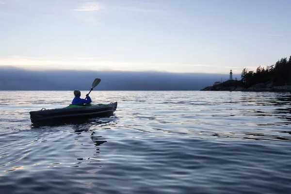 Adventurous woman is sea kayaking near a lighthouse during a vibrant and foggy winter sunset. Taken in Horseshoe Bay, West Vancouver, BC, Canada. Concept: adventure, holiday, vacation, explore