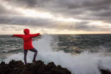 Kadın parlak bir kırmızı ceketli dalgalar kazasında bir kayalık Pasifik Okyanusu kıyısında izliyor. Alınan Ucluelet, Vancouver Adası, Bc, Kanada.