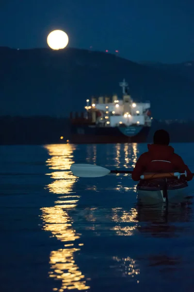 Man on a kayak watching the big moon rising over the mountain. Taken in Vancouver, BC, Canada.